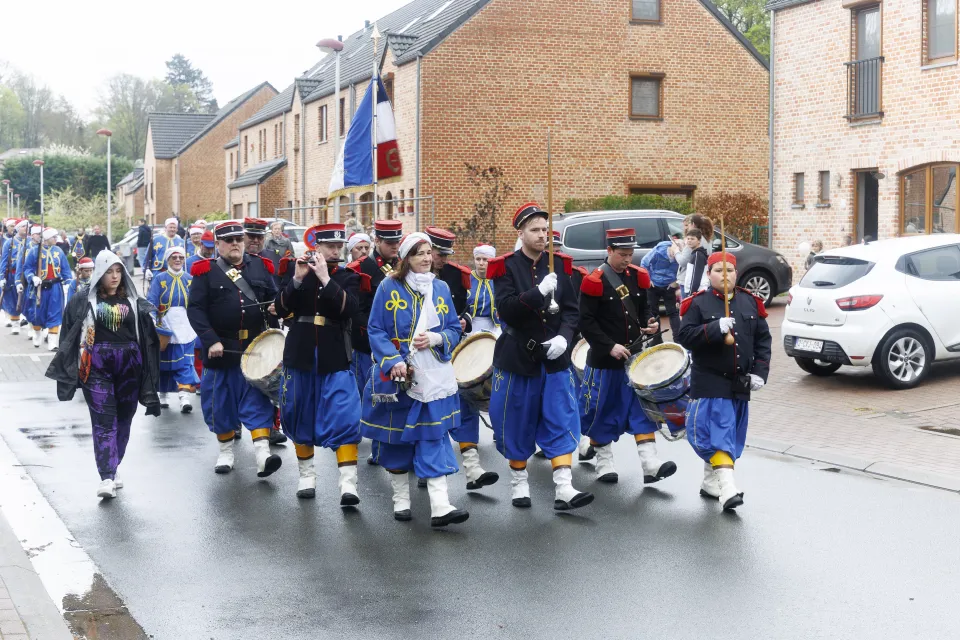Défilé folklorique à Mont-Saint-Guibert pour les 900 ans de la commune, avec une compagnie mixte en costume napoléonien bleu, rouge et noir, composée de tambours, fifres, porte-drapeau et marcheurs en habits traditionnels, avançant dans une rue résidentielle.