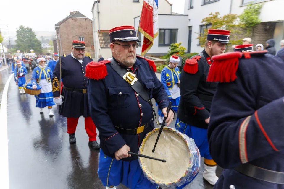 Défilé costumé à Mont-Saint-Guibert dans le cadre des 900 ans de la commune, avec des tambours et figurants en uniformes napoléoniens bleus, rouges et noirs, défilant sous la pluie dans une rue du village, accompagnés de femmes en habits folkloriques.