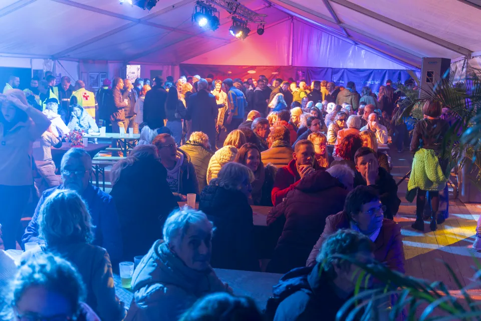 Soirée conviviale sous chapiteau à Mont-Saint-Guibert pour les 900 ans de la commune, avec des habitants attablés, échangeant dans une ambiance chaleureuse et festive, illuminée par des jeux de lumière colorés.