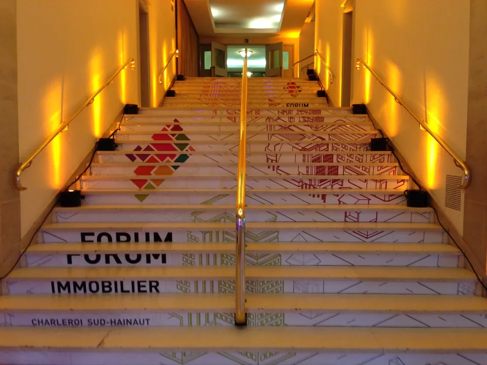 Habillage scénographique d’un escalier avec branding du Forum Immobilier Charleroi Sud-Hainaut, dans un hall d’accueil événementiel éclairé en lumière chaude."