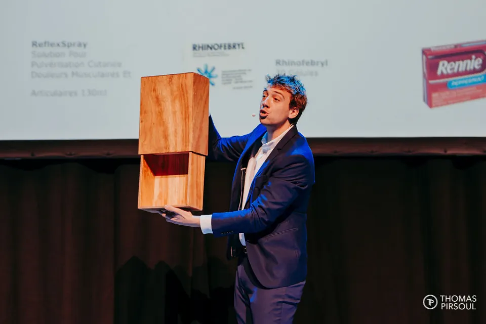 Magicien en costume bleu sur scène tenant deux cubes en bois empilés, en pleine démonstration pendant un spectacle d'entreprise