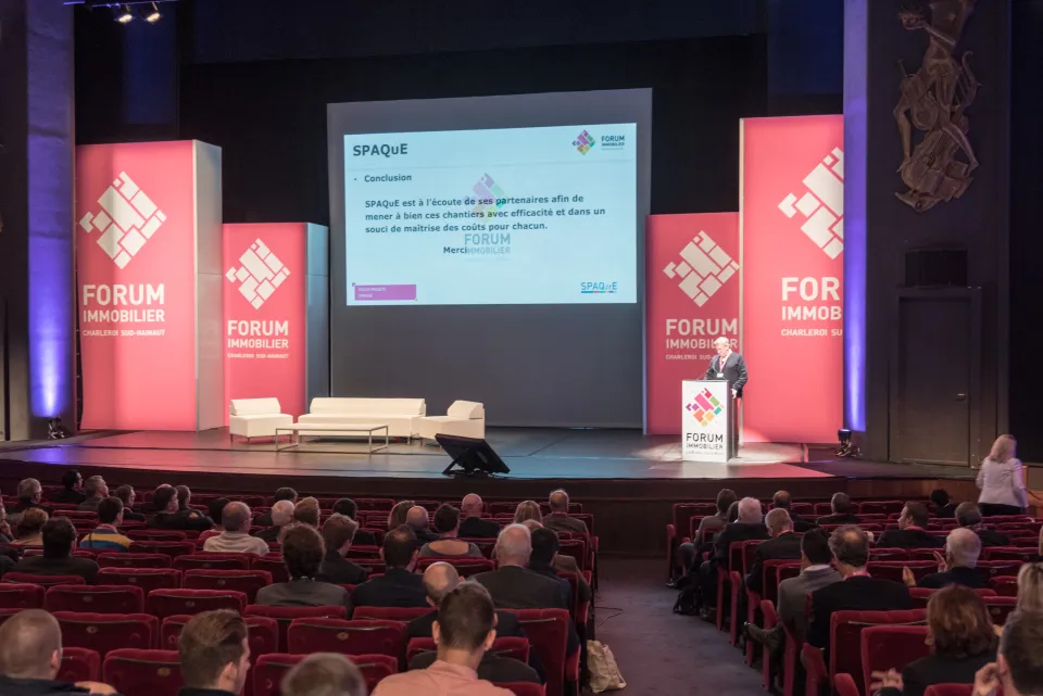 Conférence du Forum Immobilier à Charleroi Sud-Hainaut avec une présentation de SPAQuE devant un public de professionnels du secteur, dans une salle aménagée avec des panneaux de branding rose et blanc.