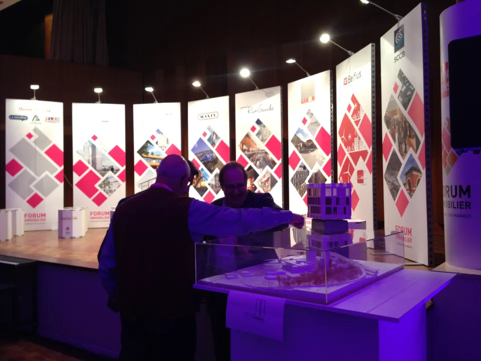 Espace partenaires du Forum Immobilier à Charleroi Sud-Hainaut, où deux participants échangent devant une maquette architecturale exposée, entourés de panneaux présentant les projets et sponsors de l'événement.