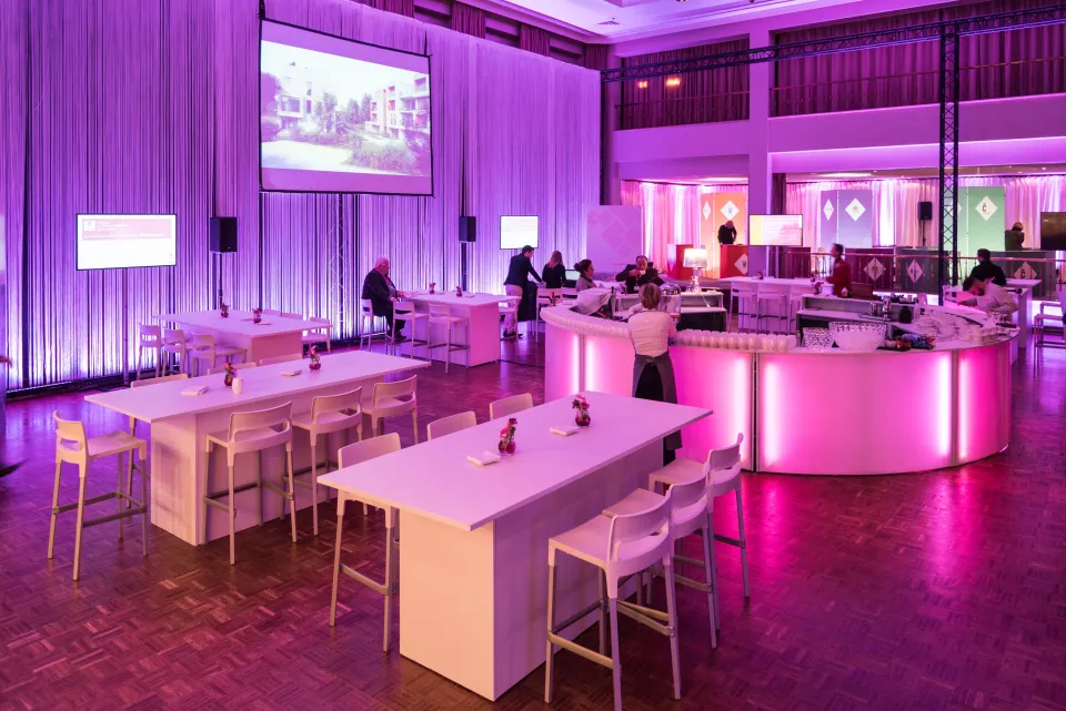 Espace networking du Forum Immobilier aménagé avec des tables hautes et un bar central, éclairé par une ambiance lumineuse rose et violette, favorisant les échanges entre les participants dans un cadre convivial et professionnel.