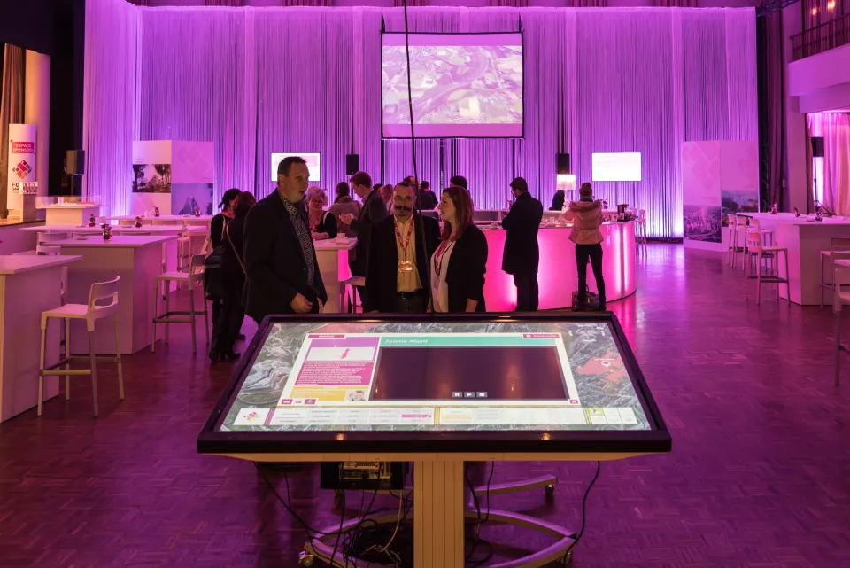 Participants en discussion autour d’un écran tactile interactif dans l’espace networking du Forum Immobilier, aménagé avec des tables hautes et un éclairage violet immersif favorisant les échanges professionnels.