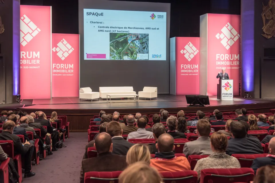 Conférence du Forum Immobilier à Charleroi Sud-Hainaut avec une présentation de SPAQuE devant un public de professionnels du secteur, dans une salle aménagée avec des panneaux de branding rose et blanc.